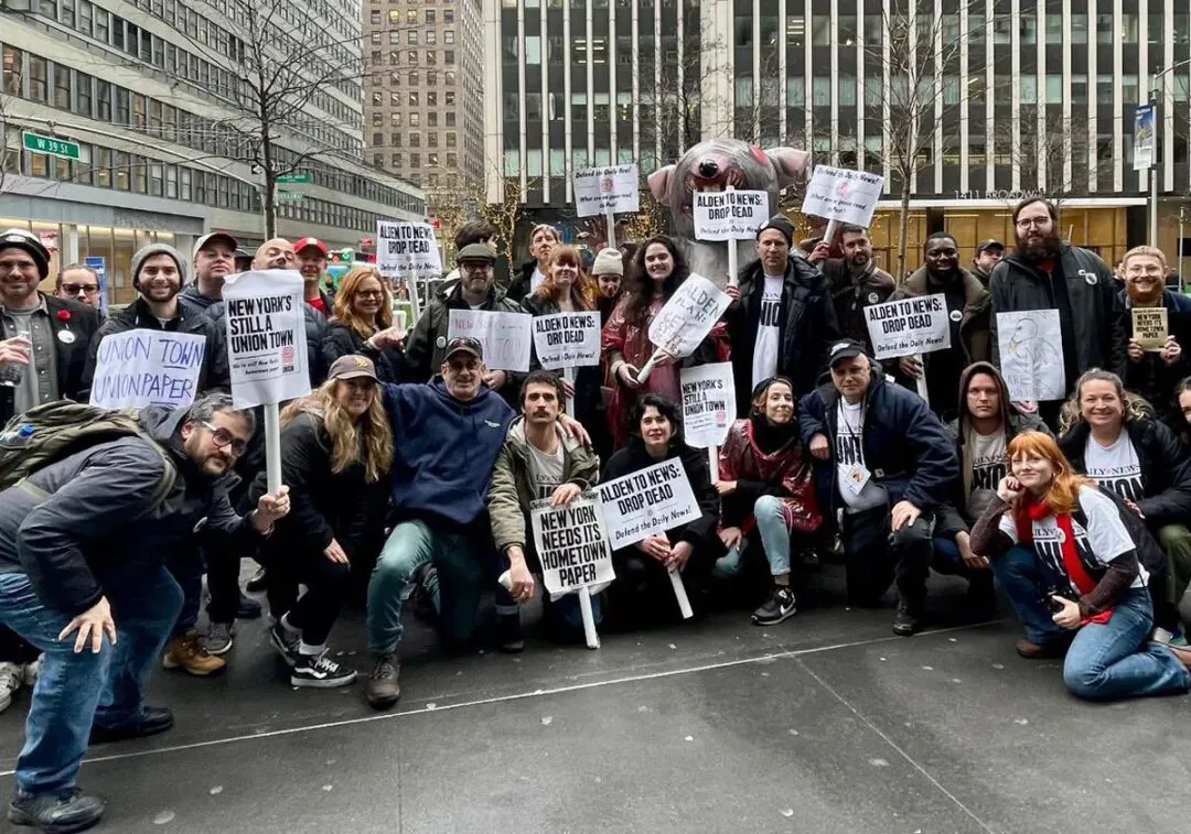 一群人在纽约街头合影，多人举着'New York's Still a Union Town''Alden to News: Drop Dead''New York Needs Its Hometown Paper'等标语牌，其中一人举着一只粉色猪猪偶。