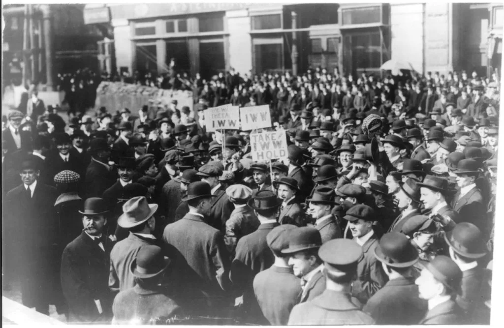 黑白老照片，1914年纽约街头聚集着密集人群，多为戴礼帽、穿西装的男性，人群中举着写有“IWW”字样的标语牌。