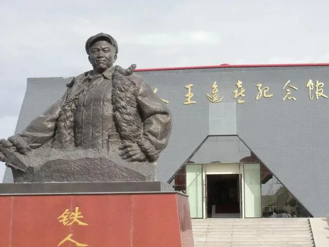 一尊头戴棉帽、身穿皮领大衣的男子铜像立于刻有“铁人”金字的红色基座上,身后是灰色纪念馆建筑,墙面有金色大字,入口呈三角形玻璃幕墙