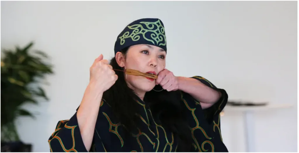身着深色阿伊努族传统长袍与头巾的女性,衣物绣有绿色漩涡纹样,双手举着竹制口簧琴凑近嘴边演奏,室内背景可见绿植。