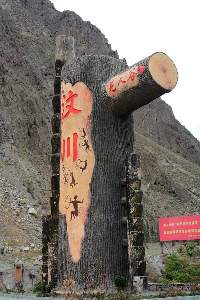 一座仿树桩造型的巨型雕塑矗立在山前，正面用红色大字写着“汶川”，上面绘有类似岩画风格的人物与动物图案，右侧断枝造型上写着“尧人谷”，背景是陡峭的山体。