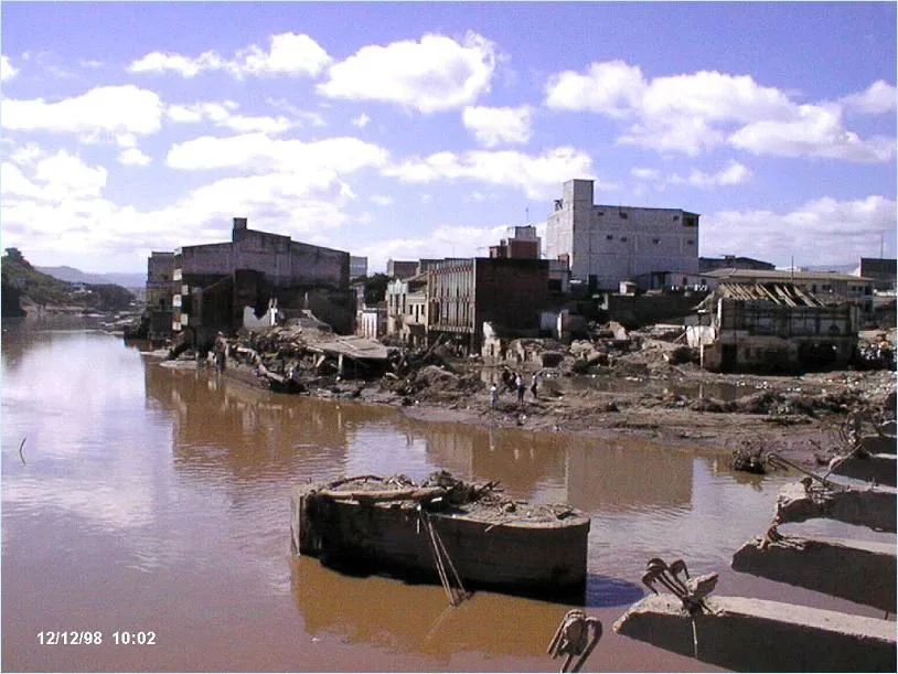 浑浊的洪水淹没河岸城区，两侧建筑严重损毁、墙体倒塌，几名人员在泥泞中查看，画面左下角有日期时间戳“12/12/98 10:02”。