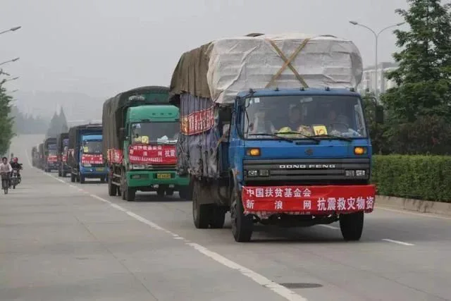 一列满载物资的卡车车队行驶在公路上，最前方卡车挂着红色横幅，写着“中国扶贫基金会 新浪网 抗震救灾物资”，路边有骑车人经过。