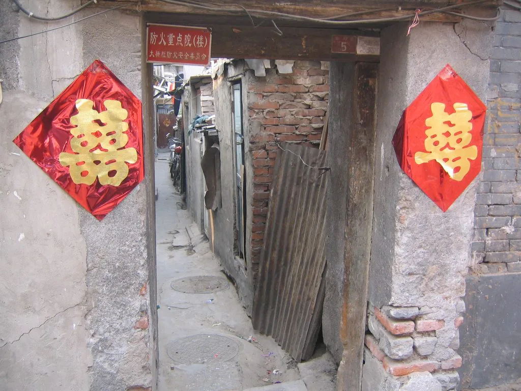 大杂院门口两侧贴着红色喜字剪纸，门框上挂着“防火重点院”标牌，门内是狭窄破旧的过道，堆放着木板等杂物。