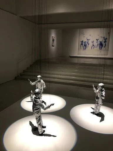昏暗展厅中，三个身穿银色金属光泽西装的人偶站在聚光灯圆圈中，头部、手部和背后延伸出黑色细绳直达天花板，背景墙上挂着一幅人物剪影画作。