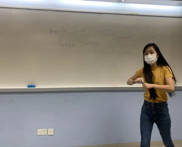 教室内，一名戴口罩、身穿黄色上衣的女子双手摆出造型动作，身后白板上手写着「Augusto Boal」「Image theatre」等字样。