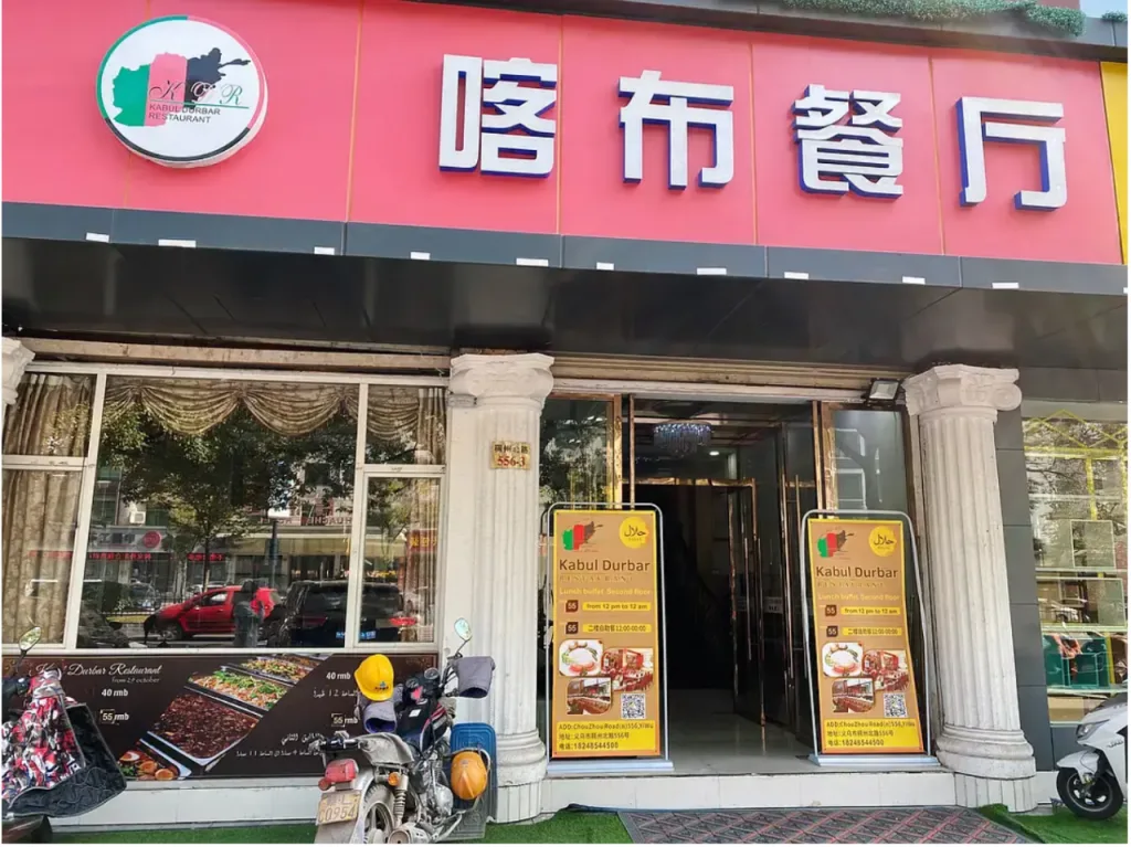 临街的阿富汗餐厅门面，招牌写着“喀布餐厅”，左侧圆形标志印有阿富汗国旗，门口立着写有“Kabul Durbar”的双语菜单牌和二维码，门前停着摩托车。