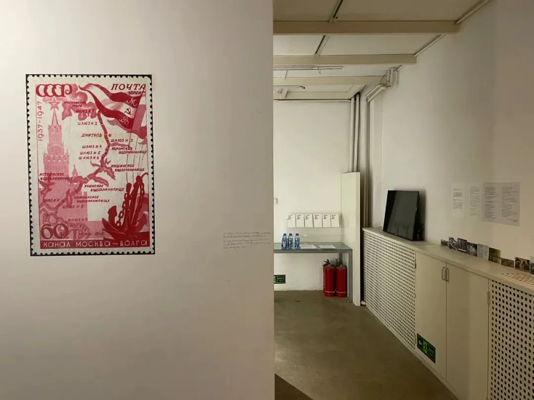 展墙左侧放大展示一张苏联红色主题邮票，印有莫斯科克里姆林宫图案、运河路线图和文字「1937-1947 运河莫斯科—伏尔加」；右侧是展厅内部的白墙走廊，墙上挂着一台平板电视和说明文字，柜台上摆放消防器材。