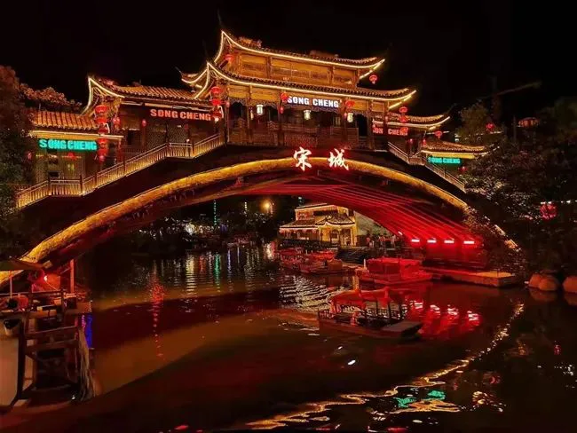 金色拱桥上悬挂“宋城”红色大字与“SONG CHENG”霓虹招牌，飞檐楼阁灯火通明，桥下河面停靠着红色游船，灯光倒映水中。