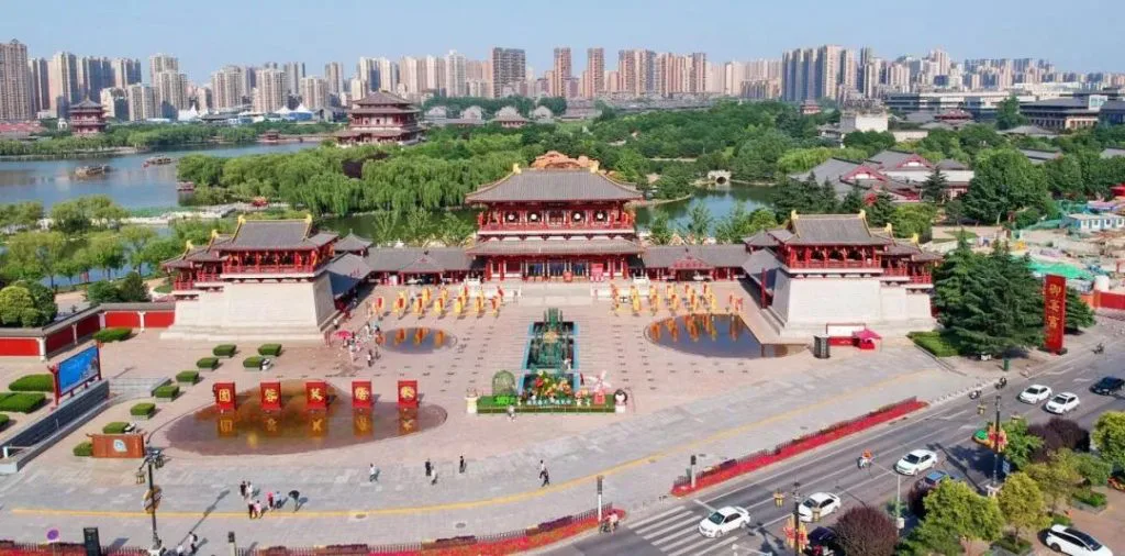 鸟瞰仿唐建筑群与广场，表演者在广场上列队表演，背景是城市高楼、湖泊与绿地。