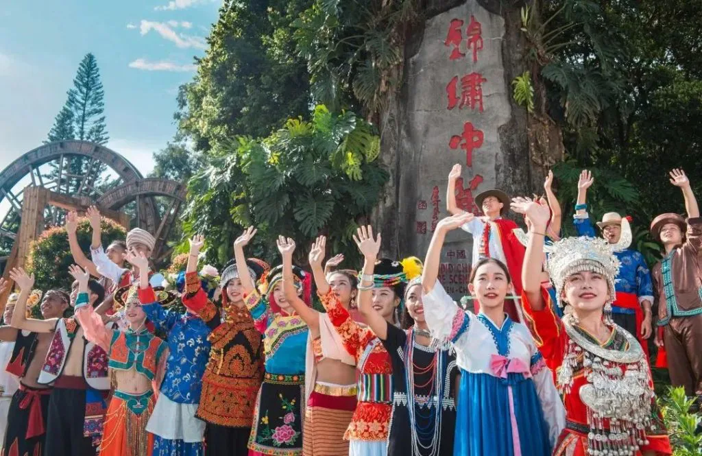 一排身着不同少数民族服饰的表演者面向镜头挥手微笑，背后是刻着“锦绣中华”字样的巨石与水车造型景观。