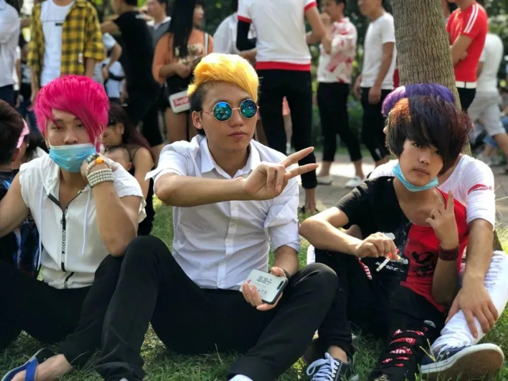 三名染着粉、黄、紫色高耸发型的青年坐在草地上，中间一人戴墨镜比出'V'字手势，两侧的人戴着口罩。