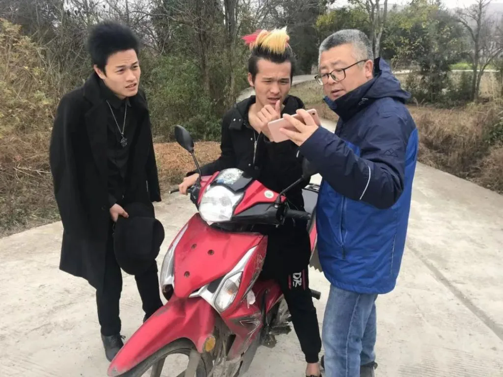 导演李一凡与两名留着夸张竖发的青年站在一辆红色摩托车旁的乡间小路上，其中一人正给他看手机。