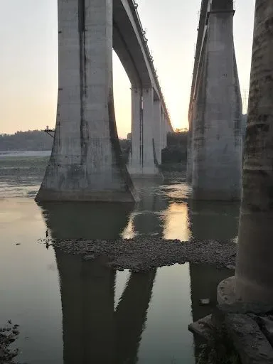 夕阳下几座高大的桥墩矗立在江面上，水中倒映出桥身，江边有一小片碎石滩，一人立于水边。