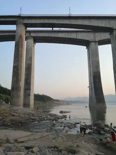 从另一角度拍摄的铁路桥墩，桥下河滩上多人戏水休憩，江面开阔，远处群山连绵。