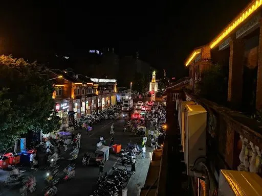 夜晚俯瞰西街，远处钟楼灯光点亮，街道两旁店铺灯火通明，路上停满摩托车，行人熙攘。