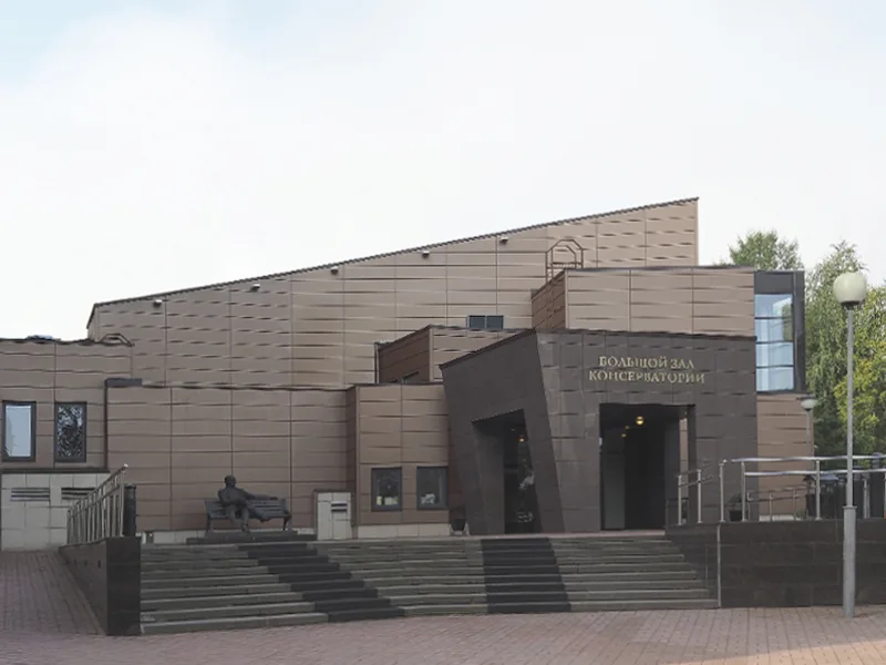 深色几何造型的音乐厅建筑外观，正门前有宽阶梯，一侧另设带扶手的坡道，台阶旁有一尊坐姿人像雕塑。