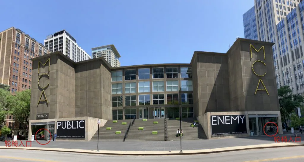 建筑正面全景，两侧塔楼贴着黄色'MCA'大字，中央宽阶梯通向玻璃门厅；图中用红圈标注两侧角落处并附文字'轮椅入口'。