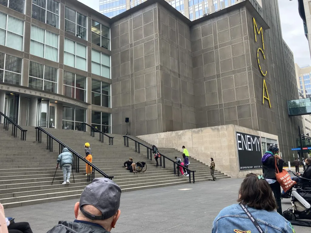 远景，MCA建筑外墙立着黄色巨型'MCA'招牌，宽阔的室外台阶上散布着穿彩色服装的表演者，前景是围观观众的背影。