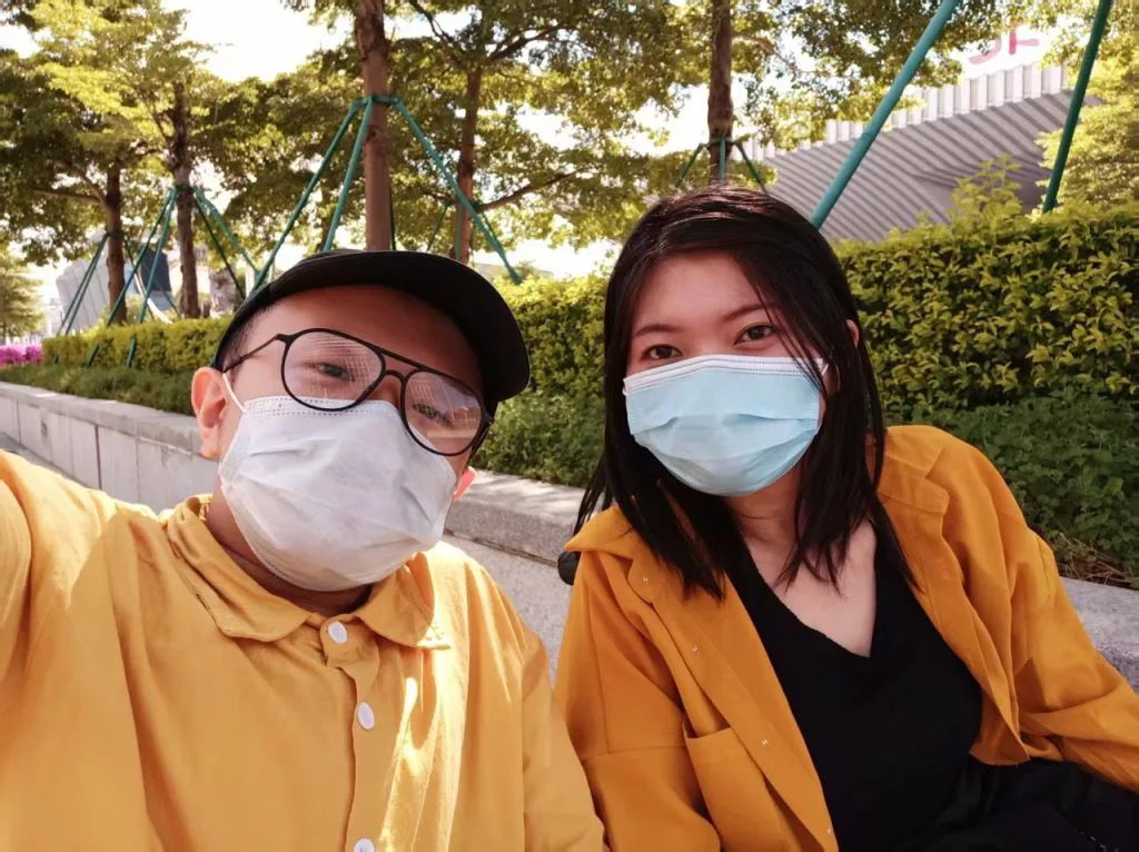 两人戴口罩并排坐在户外，一人戴鸭舌帽和眼镜，两人都穿黄色外套，身后是树丛草坪与攀爬架。