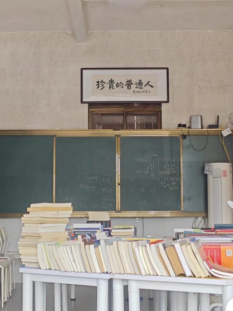 教室墙上挂着“珍贵的普通人”横幅，下方是绿色黑板，前景课桌上堆满书籍。
