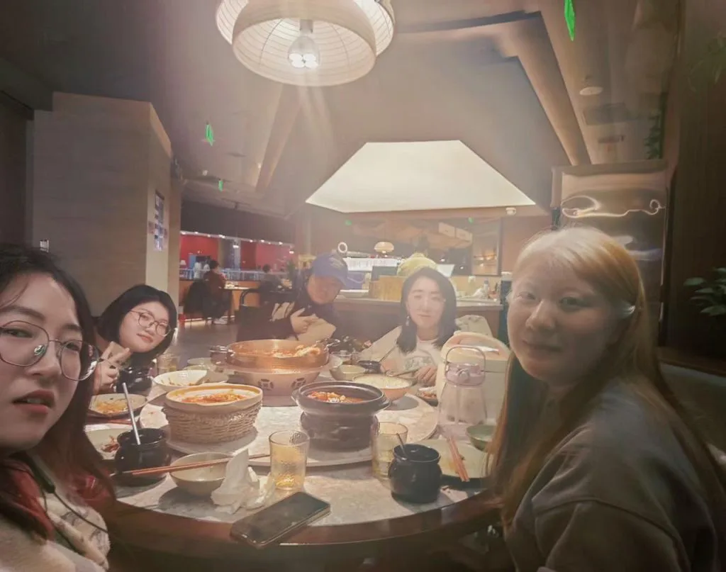 几位女性围坐圆桌用餐，桌上摆着火锅和多道菜肴，头顶吊灯洒下暖光。