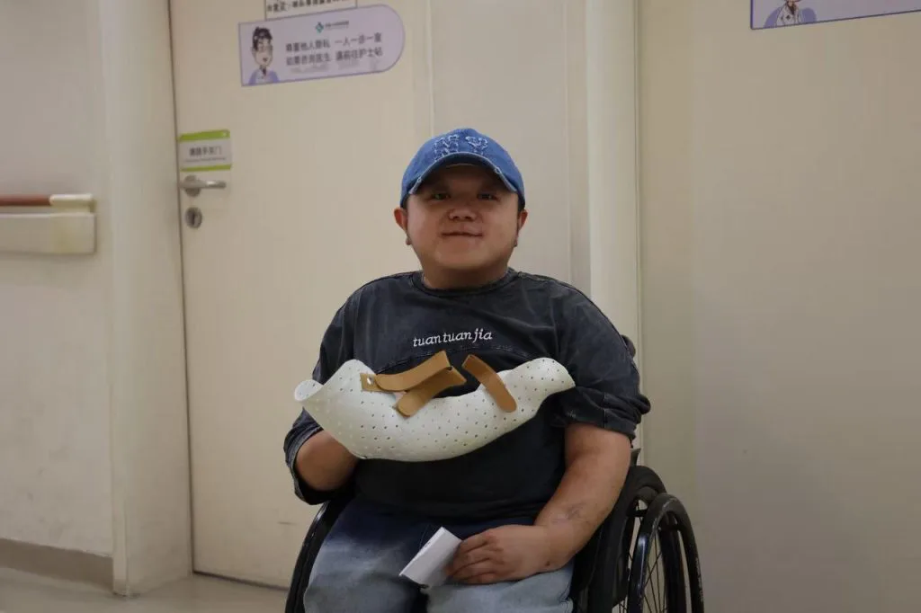男子戴蓝色棒球帽坐在轮椅上微笑，手臂戴着白色镂空支具，身后是医院走廊。