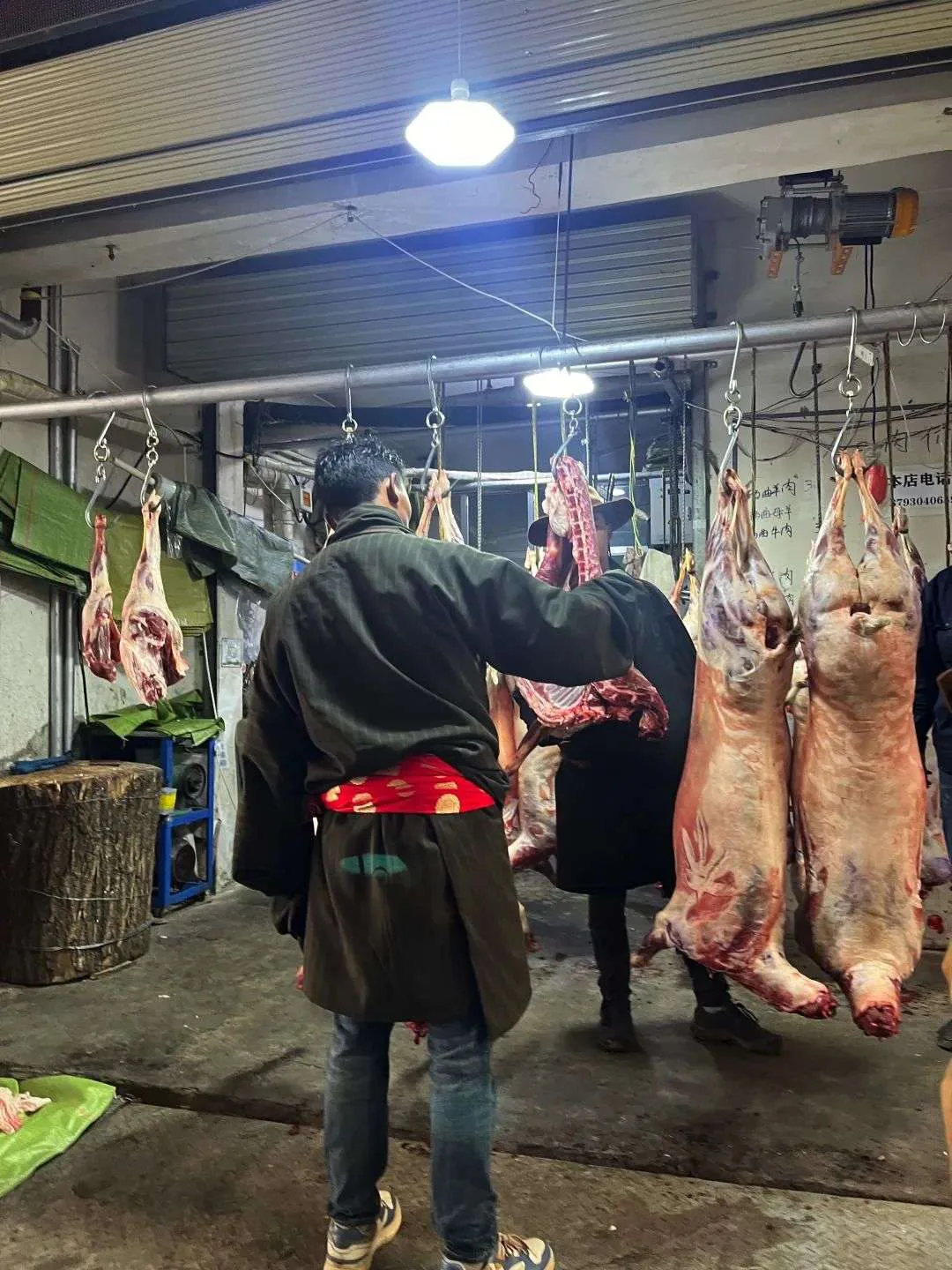 肉铺内几只整只羊胴体悬挂在铁钩上，一位穿藏式外衣的男子背对镜头站在肉架前挑选。